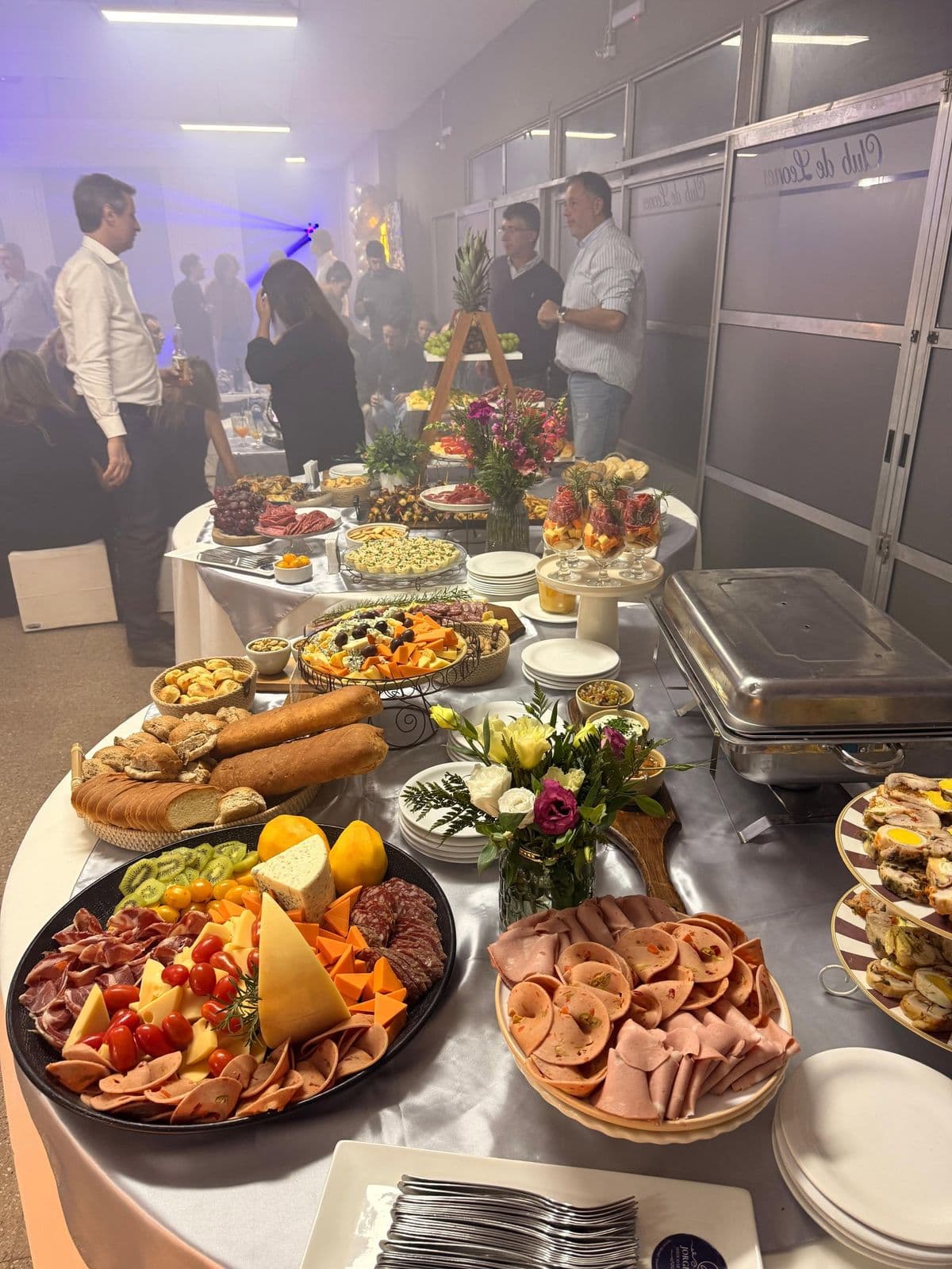 Mesa buffet en un salón con tablas de fiambres y quesos, panes, flores y fuentes