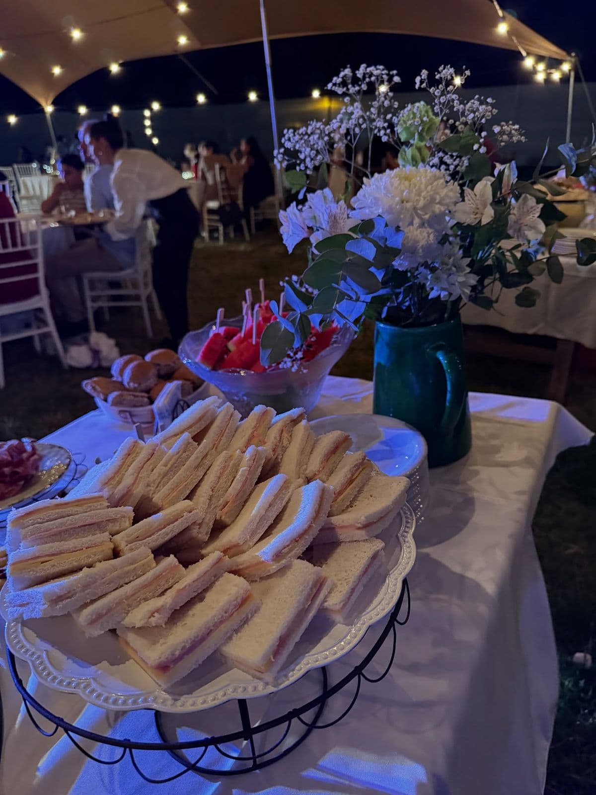 Fuente con sándwiches de miga de jamón y queso en una mesa de fiesta al aire libre, de noche