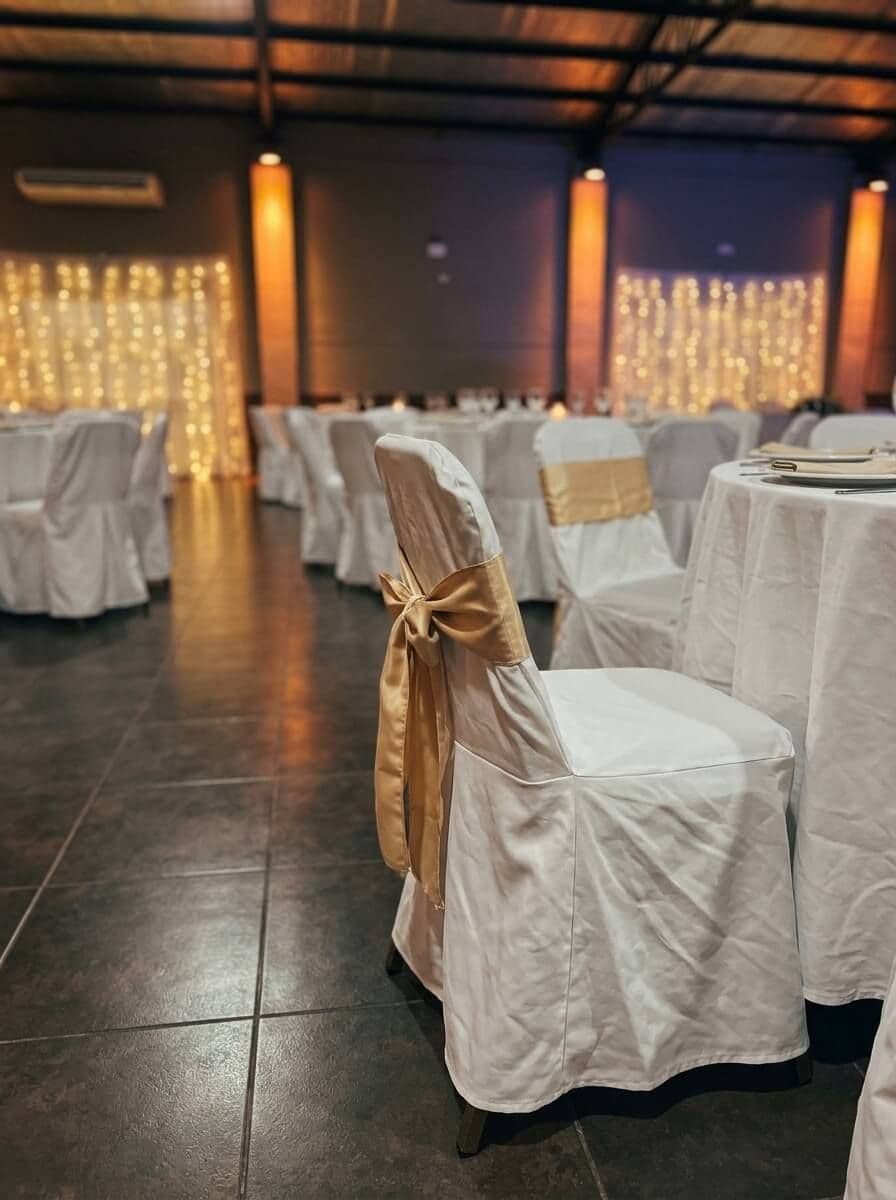 Silla con funda blanca y moño dorado junto a una mesa con mantel blanco, en un salón con luces cálidas